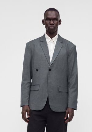 Homme à la peau foncée portant un blazer gris à double boutonnage sur une chemise blanche colletée, debout devant un fond clair uni.