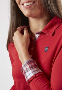Polo rouge à manches longues avec un col à carreaux, des boutons blancs et un logo brodé sur la poitrine. Tissu lisse avec une coupe décontractée.