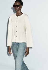 Elfenbeinfarbige, knöpfbare Jacke mit langen, weiten Ärmeln und zwei Fronttaschen, kombiniert mit hellblauen Jeans. Der Stoff hat eine weiche Textur.