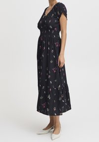 Vestido midi negro con flores, mangas cortas abullonadas, cintura fruncida, falda de capas y pequeños estampados de flores blancas y rosas. Combinado con zapatos blancos.
