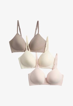 Frau mit voluminösem lockigem Haar, die einen blassrosa BH und eine blaue Jeansjacke trägt, die leicht von den Schultern rutscht, vor einem schlichten Hintergrund.