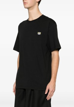 Mand iført en ensfarvet sort T-shirt med korte ærmer og et lille farverigt tegneserie-logo på venstre bryst.