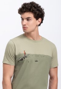 T-shirt in cotone verde oliva con una stampa grafica che rappresenta una figura arancione in piedi su delle rocce contro un paesaggio minimalista.