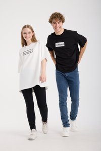 Oversized T-shirts in zwart en wit, met een rechthoekig "marat"-logo. Model combineert met zwarte leggings en spijkerbroeken, beiden met sneakers.