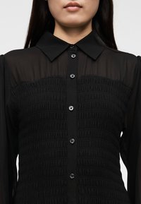 Čierna priesvitná blúzka s rozopínaním na gombíky vpredu, golierovým výstrihom a vyšívanou textúrou na hrudi s ozdobnými gombíkmi.