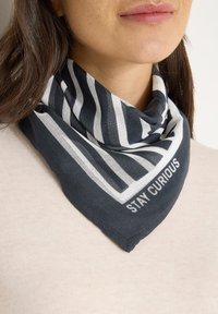 Écharpe à motifs noir et blanc avec une impression "Restez Curieux", en tissu léger, pliée en forme triangulaire, portée autour du cou.