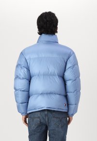 Hellblaue Steppjacke mit hochgeschlossenem Kragen, die eine glatte, glänzende Textur und ein gestepptes Design aufweist; kleines Logo in der Nähe des Saums.