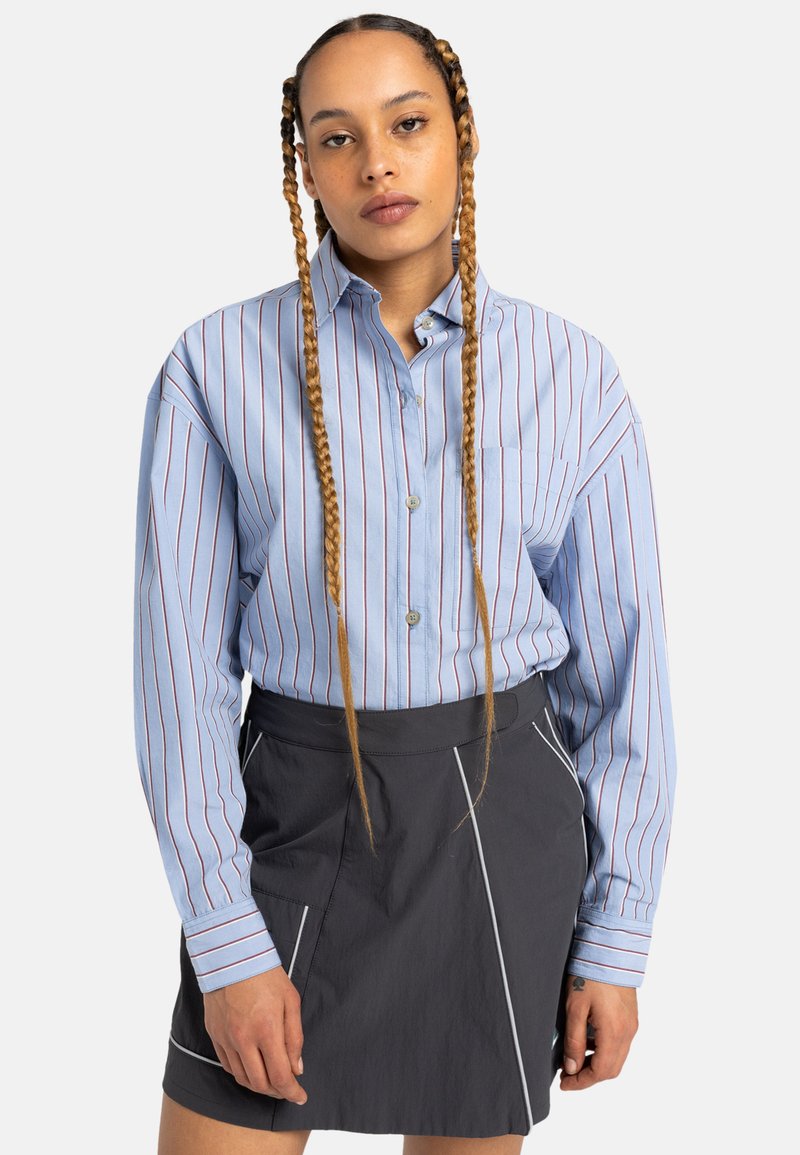 Camicia blu a righe con bottoni e maniche lunghe, abbinata a una gonna scura con piping a contrasto e vita strutturata.