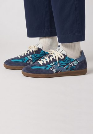 Pieds portant des baskets à motifs bleus et bleu sarcelle avec des lacets blancs, associés à des chaussettes blanches et un pantalon court bleu marine sur un fond uni.