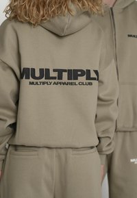 Khakiowa bluza z kapturem z odważnym czarnym napisem "MULTIPLY" oraz "MULTIPLY APPAREL CLUB" na plecach; miękka tkanina o swobodnym kroju.