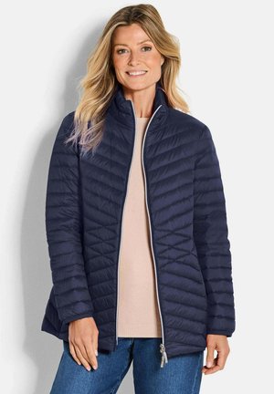 Veste matelassée bleu marine avec col haut, fermeture éclair et motif de couture horizontal. Associée à un pull léger et un jean bleu.