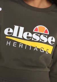 Olivgrön sweatshirt med en iögonfallande vit och gul "ellesse HERITAGE"-logotyp, med en halvkretsgrafik i röd och orange. Mjuk tygkänsla.