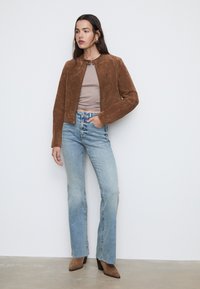 Giacca in suede marrone con scollatura rotonda e chiusura a scatto, abbinata a jeans a campana blu chiaro e stivaletti neri con tacco.