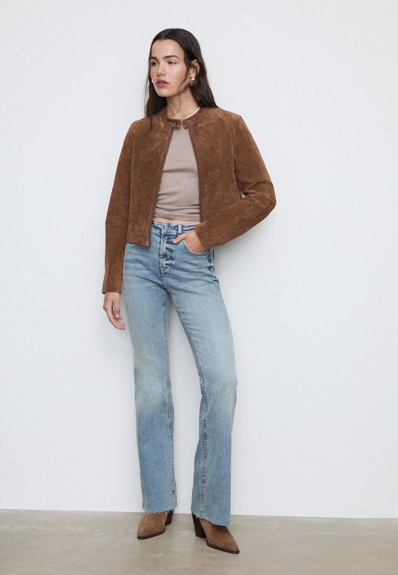 Giacca in suede marrone con scollatura rotonda e chiusura a scatto, abbinata a jeans a campana blu chiaro e stivaletti neri con tacco.