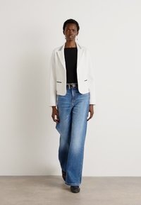 Blazer blanc avec des bordures noires, associé à un haut noir et un jean ample bleu clair. Des chaussures noires complètent la tenue.