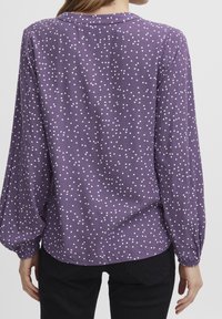 Lila blus med vita prickar, långa ärmar, mjukt tyg och rundad halsringning. Har elastiska manschetter och en avslappnad passform.