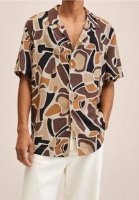 Mann trägt ein kurzärmliges Hemd mit abstrakten braunen, beigen, schwarzen und sandfarbenen Formen, kombiniert mit off-white Hosen.