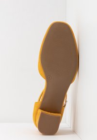 Talons en daim jaune avec une forme arrondie, texture lisse et semelle en caoutchouc dotée de rainures horizontales. Sangle réglable et détail de boucle.