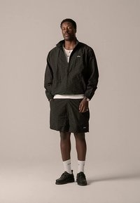 Zwarte jas met ritssluiting en bijpassende korte broek met een gestructureerd patroon. Witte T-shirt eronder, gecombineerd met zwarte schoenen en witte sokken.