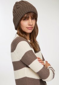 Brązowa dziana czapka typu beanie o prążkowanej fakturze i plecionym wzorze. Sweter w paski w kolorze kremowym i brązowym, z szerokimi poziomymi paskami.