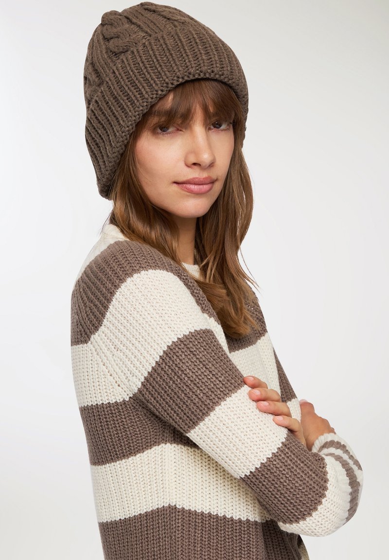 Brązowa dziana czapka typu beanie o prążkowanej fakturze i plecionym wzorze. Sweter w paski w kolorze kremowym i brązowym, z szerokimi poziomymi paskami.