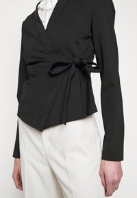 Blazer noir ceinturé avec nœud latéral porté sur une blouse blanche et un pantalon blanc, montré de la taille au cou.