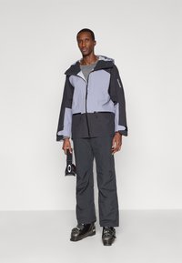 Homem escalando um padrão cinza escuro com casaco e calças de esqui, segurando óculos de esqui pretos com um logotipo branco, em pé contra um fundo claro e simples.