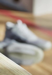 Sneakers med en tekstureret grå overdel, hvid mellemsål og unikke mønstrede snørebånd, delvist skjult mod et trægulv.