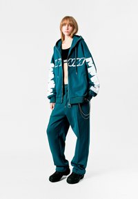 Sudadera color aguamarina con gráfico blanco en las mangas, combinada con joggers a juego. Presenta un acento de cadena plateada y calzado negro texturizado.