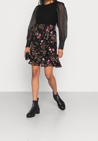 Jupe noire à fleurs avec volants et manches bouffantes en tulle transparent sur un haut ajusté ; associée à des bottines noires et tenant un petit sac noir.