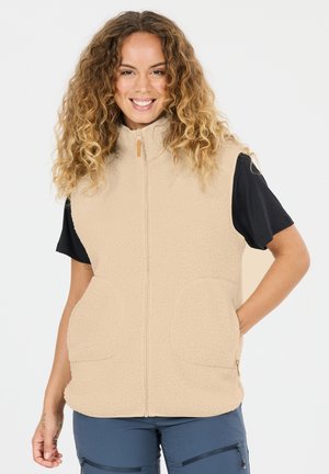 Lächelnde Frau mit lockigem Haar, die eine beige Fleeceweste über einem schwarzen Kurzarmshirt und blaue Hosen mit Reißverschluss trägt, Hände in den Westentaschen.