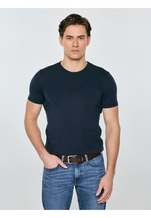 Mladić s kratkom smeđom kosom, obučen u prilagođenu tamnoplavu majicu kratkih rukava, plave traperice i smeđi remen, stoji ispred jednobojne svijetle pozadine.