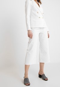 Bílé dvouřadé sako se zlatými knoflíky, spojené s bílými zkrácenými džíny a černými texturovanými sandály na blokovém podpatku.