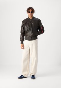 Casaco bomber de couro castanho com punhos e colarinho canelados, combinado com calças largas em creme e ténis azul marinho com riscas em azul claro.