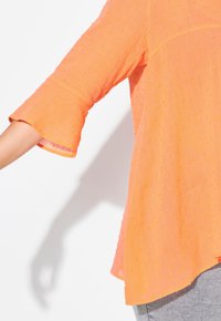 Blusa arancione con texture, maniche corte a trombetta e piccoli pois gialli. Presenta una vestibilità ampia e un colletto rotondo.