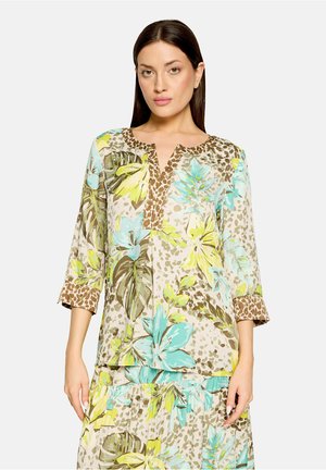 Frau trägt eine Bluse mit Blumenmuster in Türkis, Gelb und Grün sowie Leopardenmuster-Akzenten am Ausschnitt und an den Ärmeln.