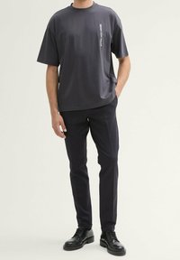 Mörkgrå t-shirt med kort ärm och rak skärning, med "DENIM TOM TAILOR" tryckt vertikalt i vitt. Matchas med marinblå byxor och svarta skor.