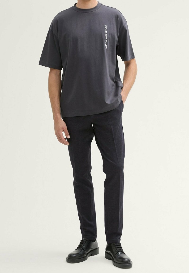 Mörkgrå t-shirt med kort ärm och rak skärning, med "DENIM TOM TAILOR" tryckt vertikalt i vitt. Matchas med marinblå byxor och svarta skor.