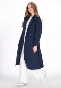Marineblauwe trenchcoat gemaakt van gladde stof, met een dubbelrijig ontwerp, gouden knopen en zijopeningen. Draagt over een wit outfit en sneakers.