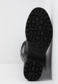 Semelle de botte noire avec un motif de grip texturé, affichant le nom de la marque Tamaris et la taille 37, inclinée contre un fond blanc.