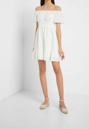 Mujer con vestido blanco de hombros descubiertos, bajo con volantes y mangas cortas, combinado con sandalias de esparto beige en un estudio sencillo.