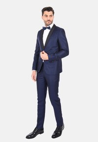 Tuxedo blu navy con colletto in satin nero, pantaloni coordinati e una camicia bianca. Presenta un papillon nero e scarpe lucide nere.
