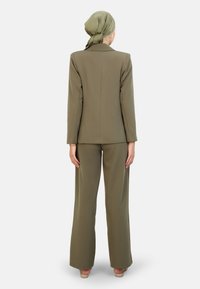 Costume ajusté vert olive avec manches longues, pantalons à jambes larges et texture lisse, associé à un foulard assorti et des accessoires minimaux.