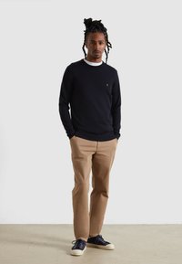 Marineblauer Rippstrickpullover mit Rundhalsausschnitt, beige Chinos und blauen Sneakers mit weißen Sohlen. Das Model hat drehendes Haar und steht vor einem neutralen Hintergrund.