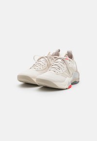 Light beige athletic shoes made from textured mesh and synthetic materials, featuring white laces and contrasting grey and red accents.