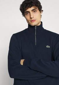 Tamsiai mėlyna zip-up sportinė striukė su aukštu apykakle, tekstūra su raukšlėmis ir Lacoste krokodilo logotipu ant kairiojo krūtinės šono. Ilgos rankovės, laisvas kirpimas.