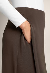 Gonna marrone con tasca laterale, abbinata a una maglietta a maniche lunghe bianca a costine. Sono visibili una texture liscia e una silhouette snella.