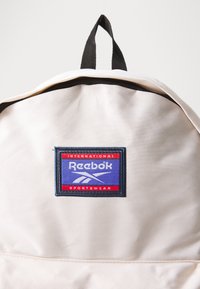 Cremfarbener Canvas-Rucksack mit schwarzem Reißverschluss und Griff. Auf der Vorderseite befindet sich ein rechteckiges, farbenfrohes Reebok-Logo-Patch.