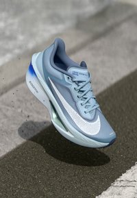 Světle modrá běžecká obuv Nike Zoom Fly s bílým swooshem a podrážkou ZoomX, která levituje nad asfaltovou silnicí poblíž bílé pruhy.