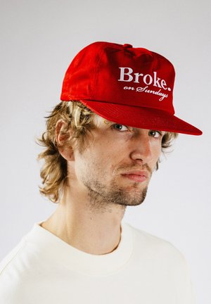 Junger Mann mit hellbraunen lockigen Haaren, der eine rote Mütze trägt, auf der "Broke. on Sundays" steht, und ein weißes Rundhalsshirt vor einem hellen Hintergrund.